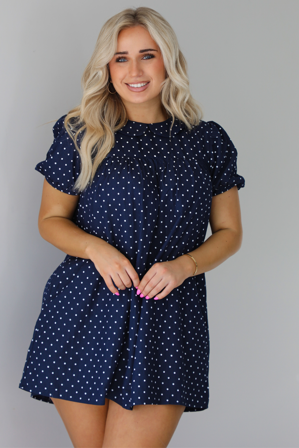 Woman wearing a navy baby-doll cut dress with a white polkadot print pattern throughout. The dress has puffed short sleeves and functional pockets on either side. The photo is taken indoors against a white wall.