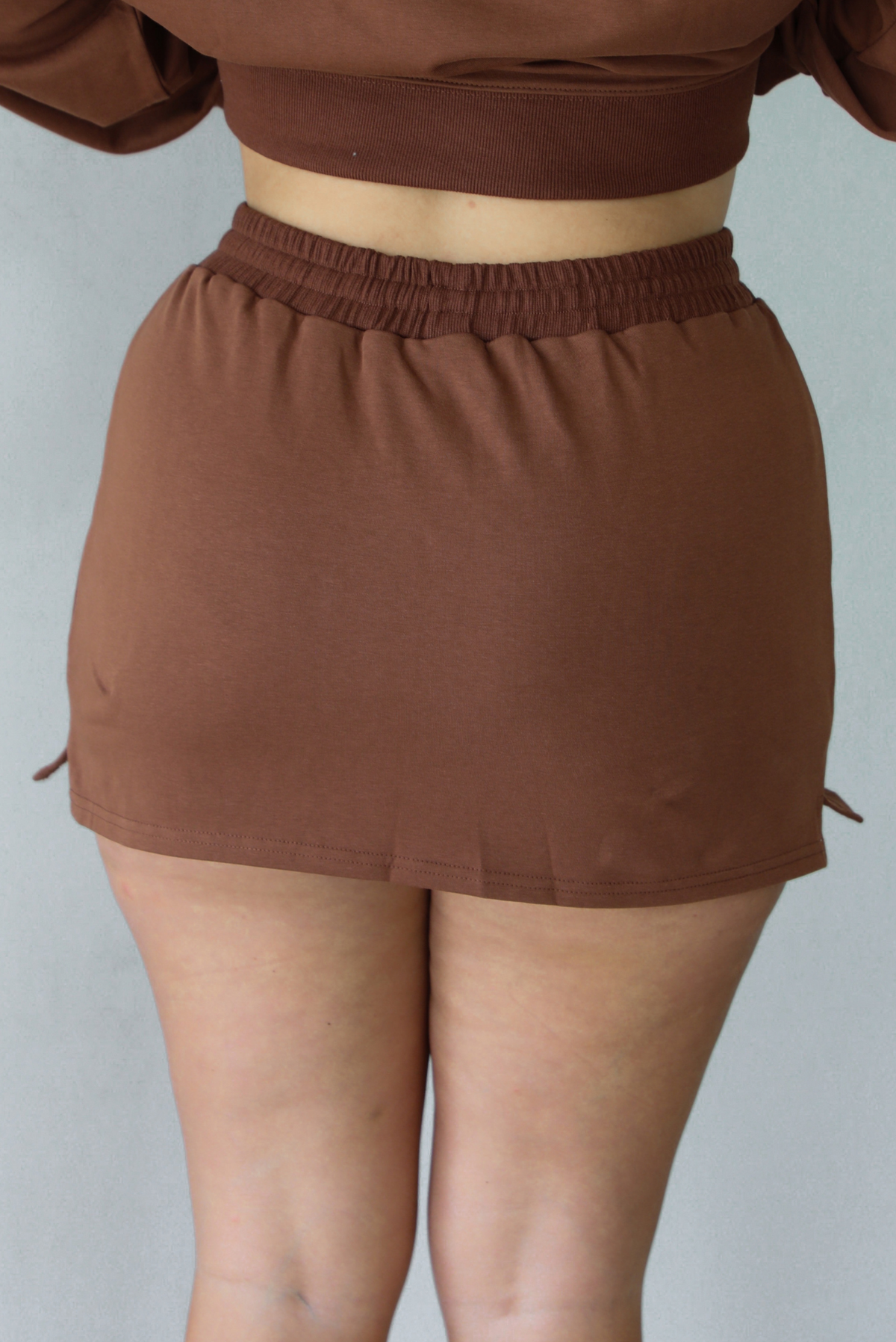 A woman shown from behind wearing a brown matching two piece set featuring a cropped sweatshirt and mini skirt of the same material, standing indoors against a white wall. The photo shows a closeup of the back of the skirt. 