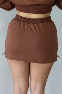 A woman shown from behind wearing a brown matching two piece set featuring a cropped sweatshirt and mini skirt of the same material, standing indoors against a white wall. The photo shows a closeup of the back of the skirt. 