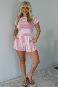 Woman wearing a pink and multi-colored striped patterned two piece set featuring a cropped t-shirt and high waisted, elastic waist shorts indoors against a white wall with white marble flooring. 