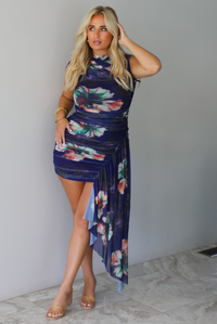 A blonde model wearing a sleeveless, high-neck mini dress with a navy blue base and a colorful floral print, standing indoors against a white wall with white marble flooring. 