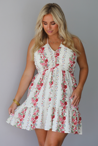 A woman wearing a white halter mini dress with a red and pink floral print, standing indoors against a white wall.