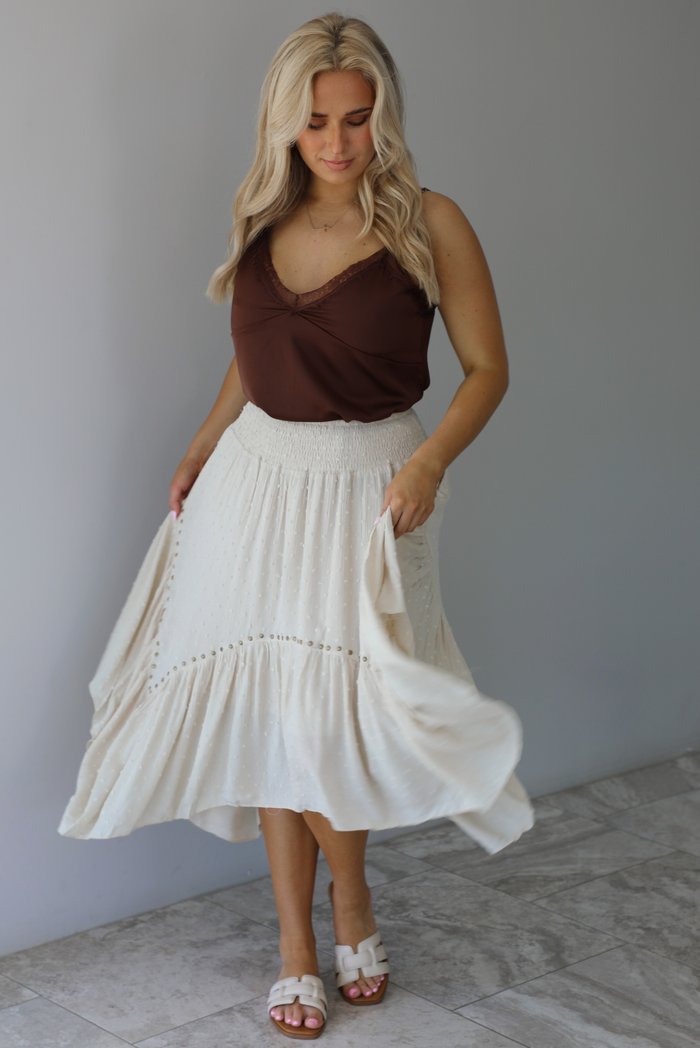 Woman wearing a satin brown tank that is tucked into a high waisted sand colored midi length skirt. The photo is taken in front of a white wall with white marble flooring. 