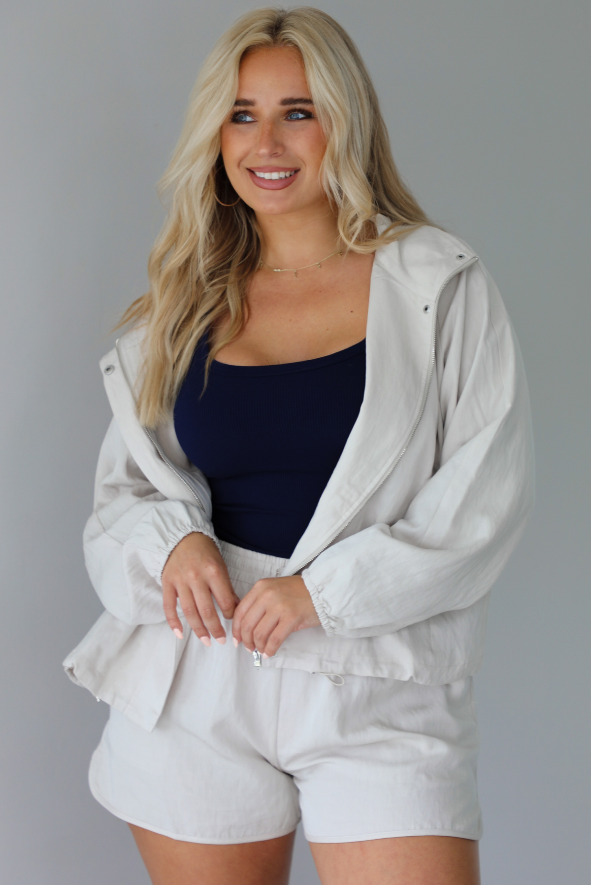 Woman wearing a stone colored two piece set featuring a zip up jacket and shorts with a navy tank tucked into the shorts indoors against a white wall.