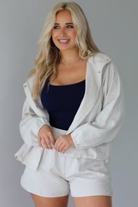Woman wearing a stone colored two piece set featuring a zip up jacket and shorts with a navy tank tucked into the shorts indoors against a white wall.