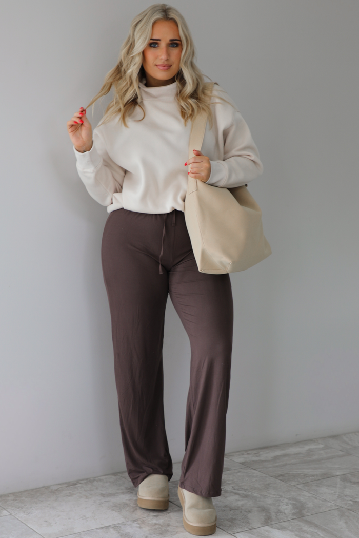 Woman wearing a beige high-neck, oversized sweater, high waisted brown pants, with a beige shoulder bag, and beige ugg boot shoes. The photo is taken indoors against a white wall with white marbled flooring. 