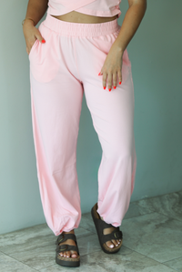 Woman wearing a pink two piece set featuring a cropped short sleeved top and high waisted jogger-style pants indoors against a white wall with white marble flooring. The photo shows a closeup of the front of the pants, which have an elastic waistline, functional pockets on either side, and a cinched detail at the ankles. 