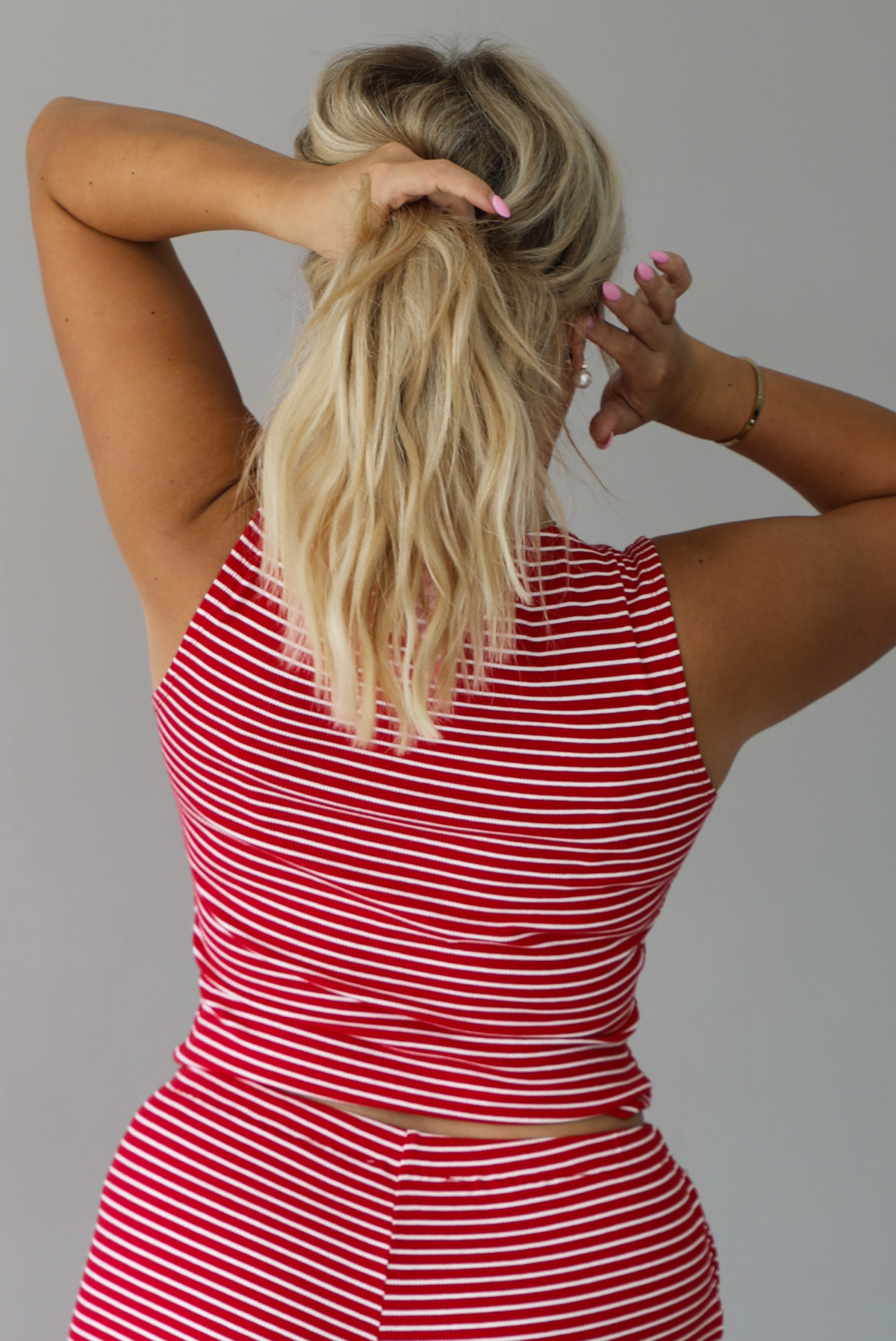 A woman modeling a red and white striped sleeveless set consisting of a tank top with non-functional button closures in the front and high waisted pants with a drawstring waistline and pockets on either side indoors against a white wall. The photo shows a close-up of the back of the tank.