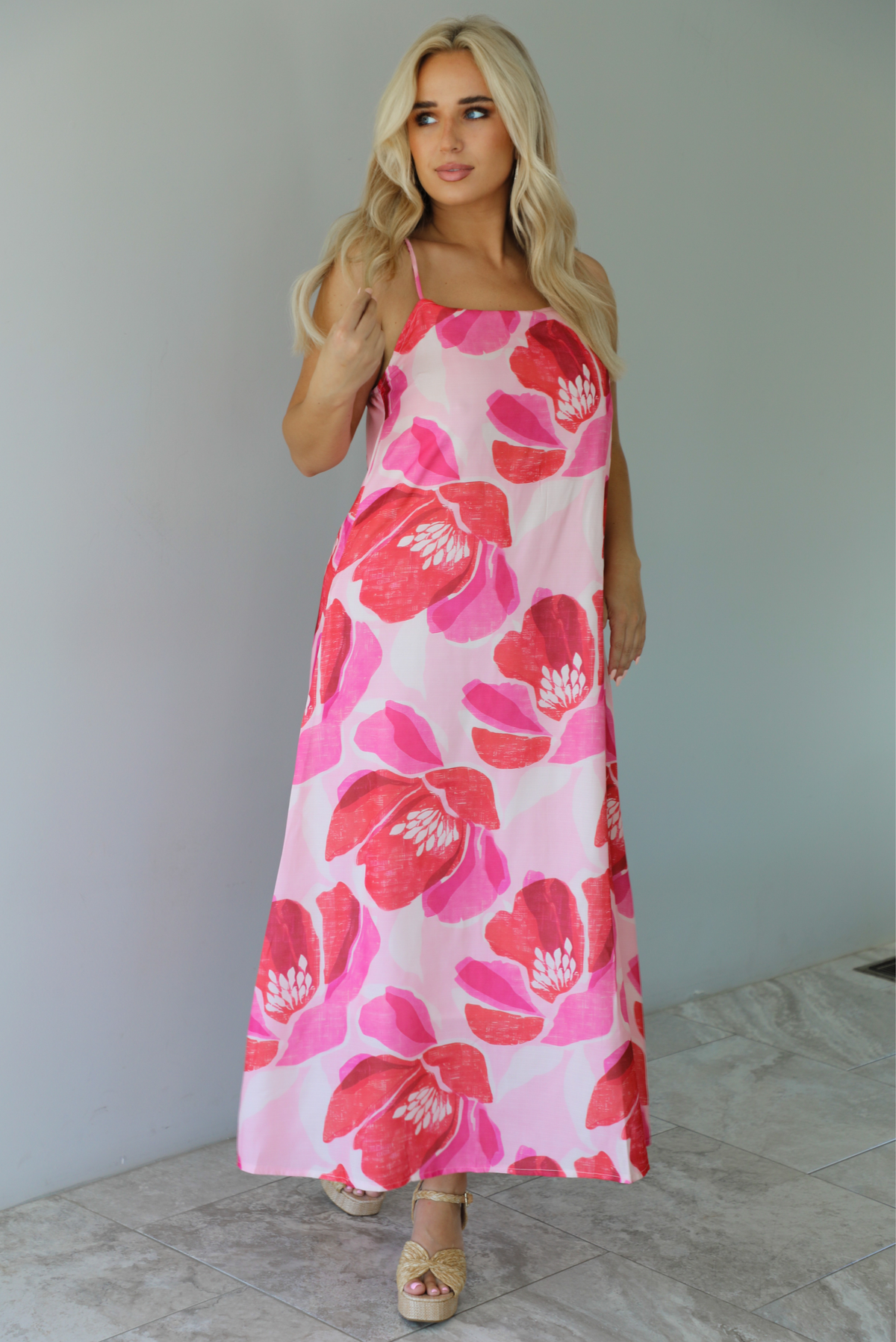 Woman wearing a pink and red floral print maxi dress with thin straps and a flowy fit indoors against a white wall with white marble flooring. 