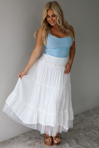 Woman wearing a blue tank and high waisted white maxi-length skirt indoors against a white wall with white marble flooring. 