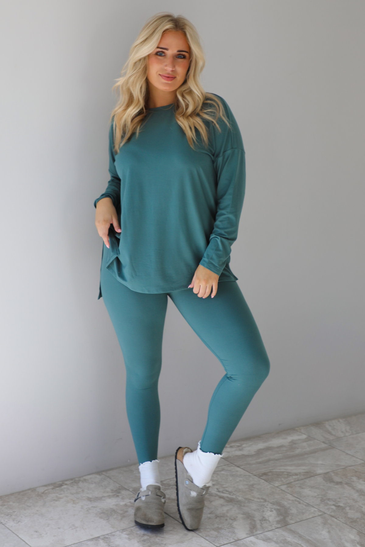 Woman wearing a teal two piece set that features an oversized long sleeved top and fitted leggings. The set is paired with high ankle socks and grey slide on shoes. The photo is taken indoors against a white wall with white marble flooring.