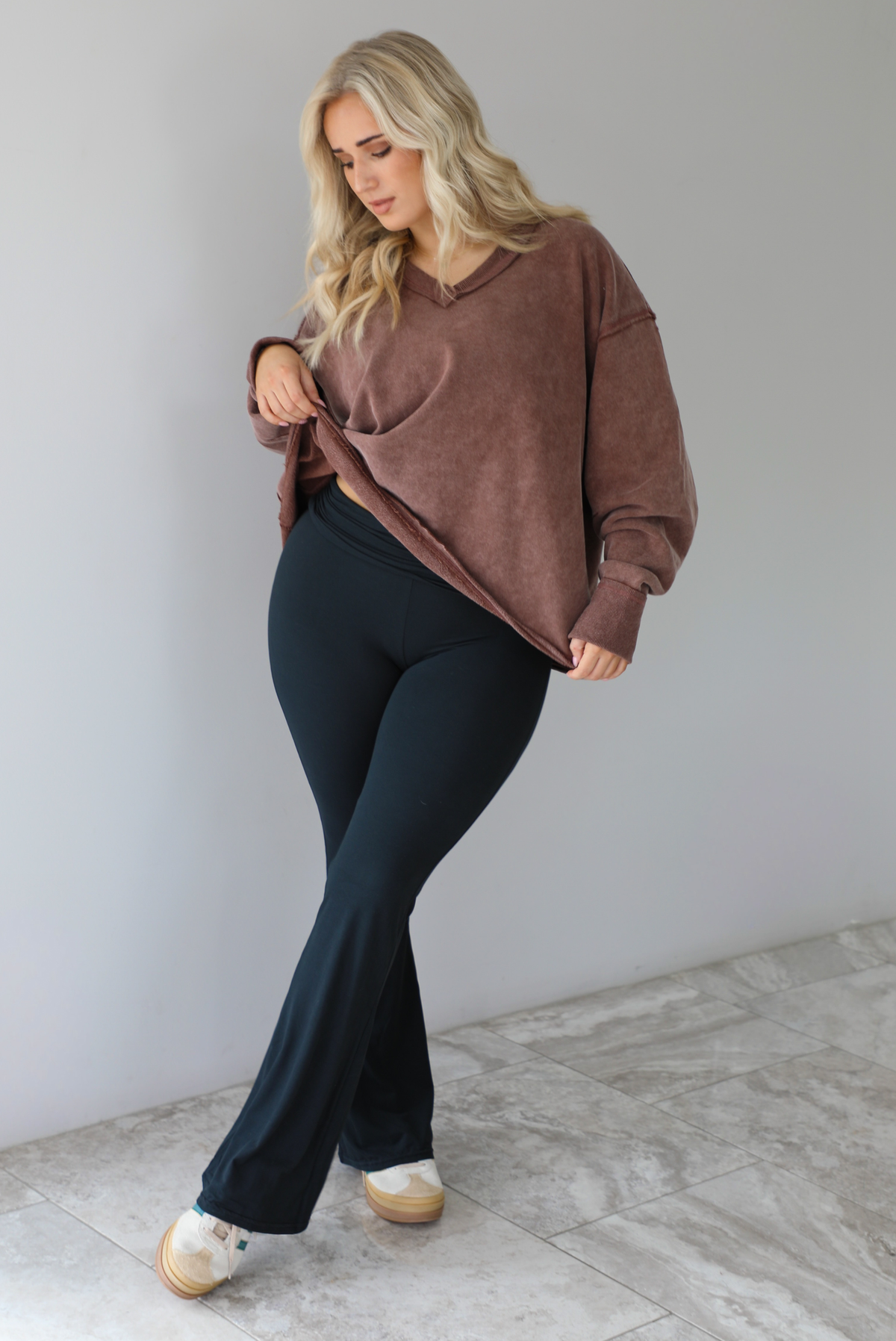 Woman wearing a mauve oversized v-neck sweater with raw hem details with high waisted black flared leggings. The photo is taken in front of a white wall with white marble flooring. 