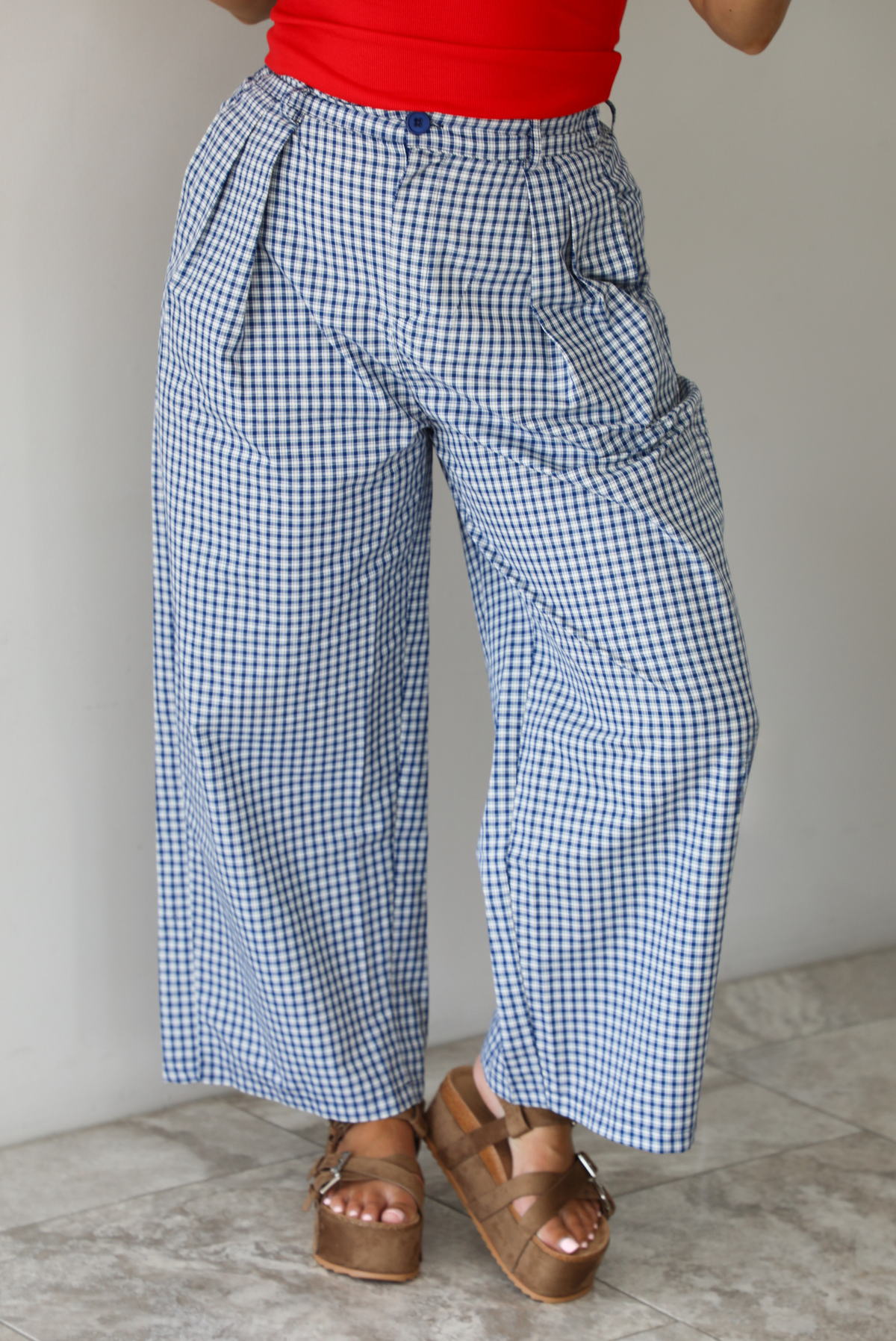Woman wearing a navy and white plaid print pants that have a high waist, pleated details, functional pockets on either side, and a professional fit indoors against a white wall with white marble flooring. 