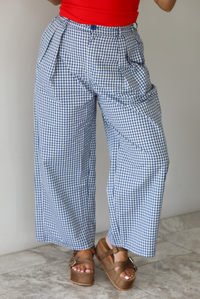 Woman wearing a navy and white plaid print pants that have a high waist, pleated details, functional pockets on either side, and a professional fit indoors against a white wall with white marble flooring. 