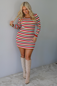 A woman wearing a red, blue, white, and brown striped long sleeved mini dress, paired with sand colored boots, standing indoors against a white wall with white marble flooring. 