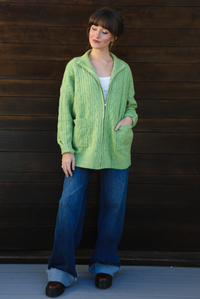 Woman model wearing a green cable knit zip-up cardigan with dark denim jeans outdoors against a dark brown wooden door. 