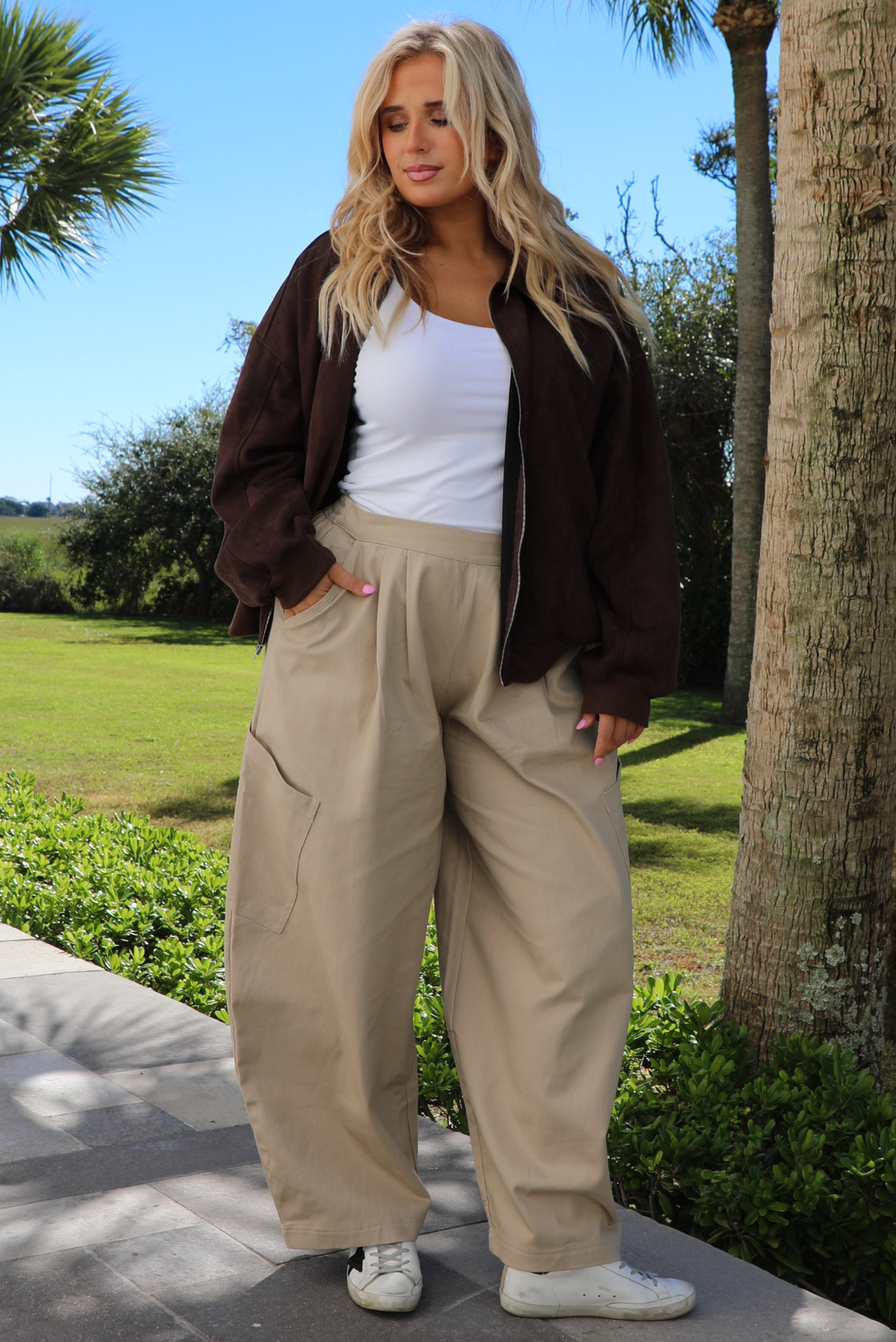 A woman wearing a brown suede jacket with a white tank and sand colored baggy pants, standing outdoors on a stone path with palm trees and other greenery in the background. 