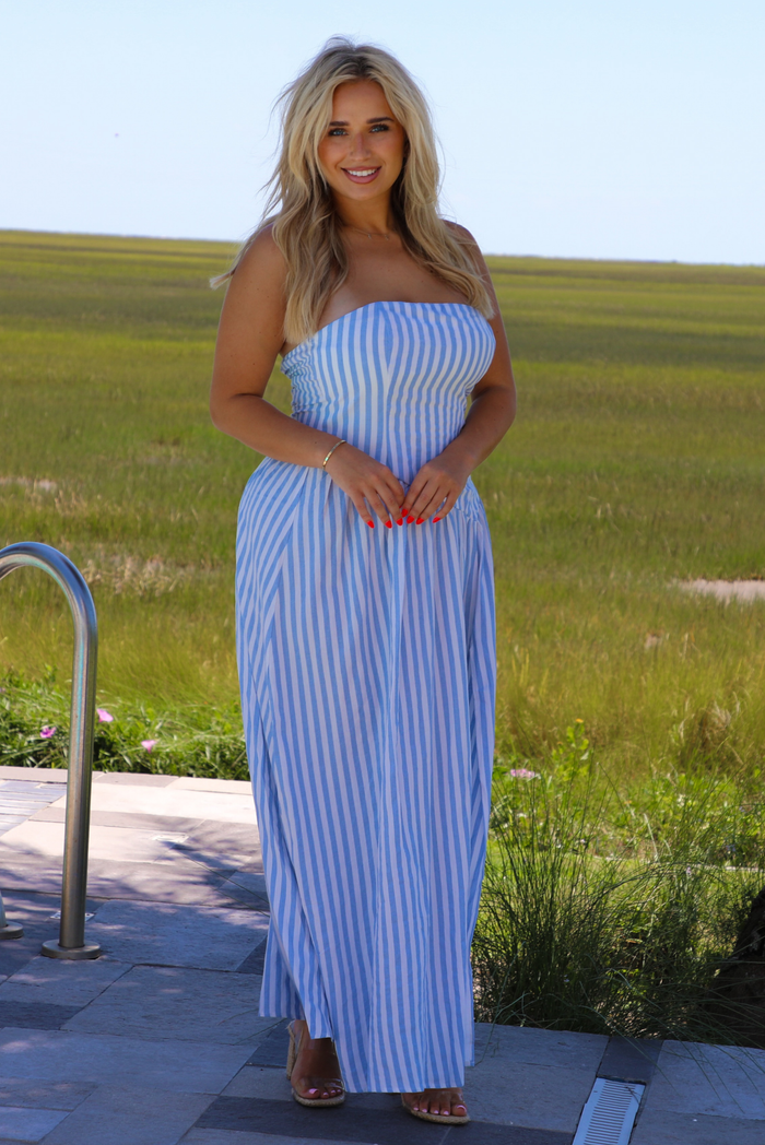 Woman wearing a blue and white striped strapless maxi dress outdoors while standing on a sidewalk with greenery in the background.