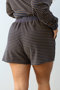 Close up of a woman model wearing a brown and multi-colored striped pattern throughout. The set consists of an oversized top with quarter-button details and shorts. The photo is taken outdoors against a white wall. The photo showcases the back of the shorts that have an elastic and drawstring waistline with functional pockets on either side.