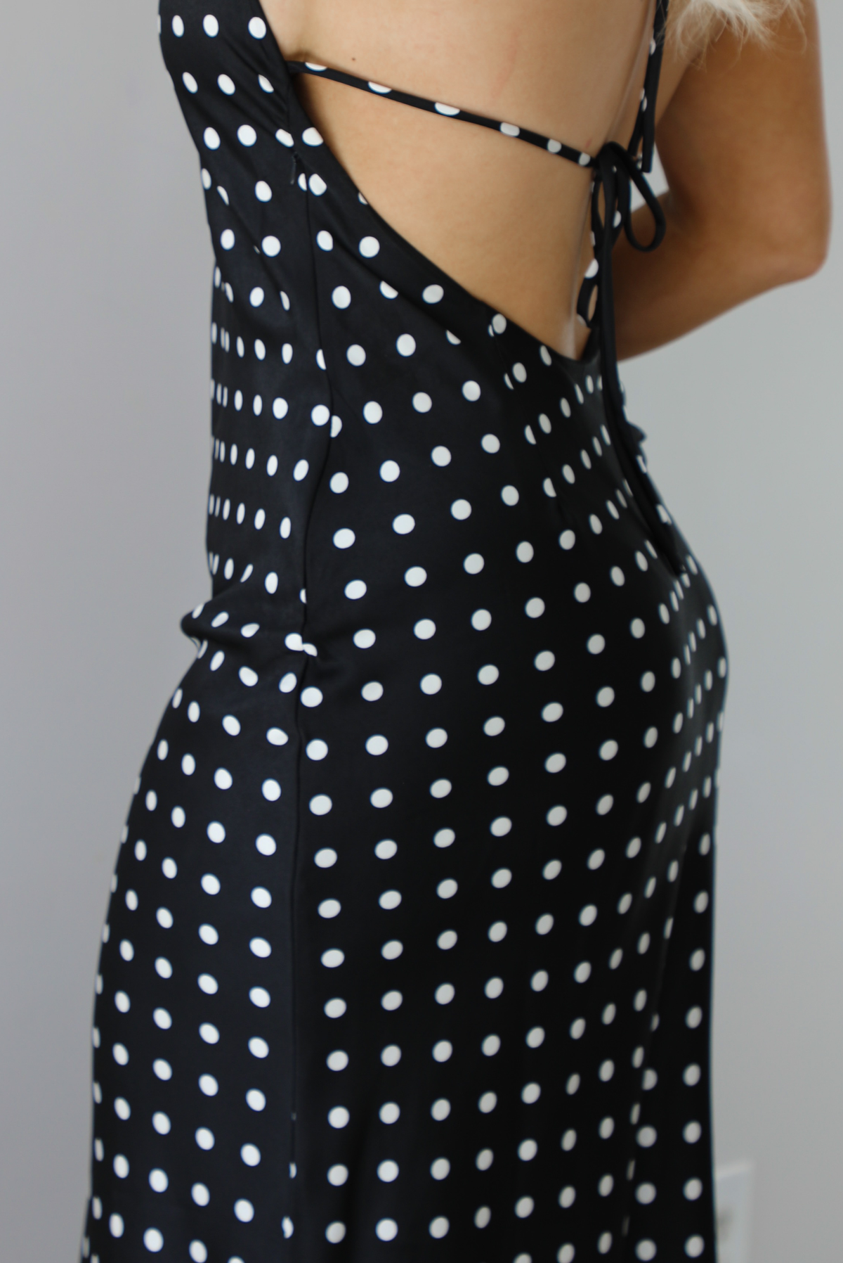 Woman wearing a black wearing a white polka dot printed midi dress that has a halter tie neckline, is form fitting, with white lace trim at the bottom. The photo is taken indoors against a white wall and shows the side of the dress with the zipper closure.