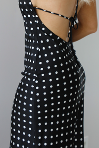 Woman wearing a black wearing a white polka dot printed midi dress that has a halter tie neckline, is form fitting, with white lace trim at the bottom. The photo is taken indoors against a white wall and shows the side of the dress with the zipper closure.