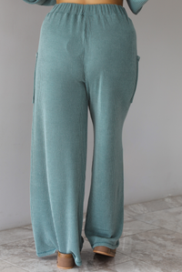 Woman wearing a teal two piece set that has a v-neck long sleeved oversized top and high waisted pants. The photo shows a closeup of the back of the pants of the set, which have an elastic waistline, functional pockets on either side, with a ribbed texture.  The photo is taken indoors against a white wall with white marble flooring. 