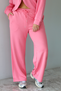 Woman wearing a pink sweat-set featuring a cropped zip-up hoodie with pants.  The pants have an elastic and drawstring waistline, functional  pockets, and a relaxed fit. The photo shows a closeup of the pants. The photo is taken indoors against a white wall with white marble flooring. 