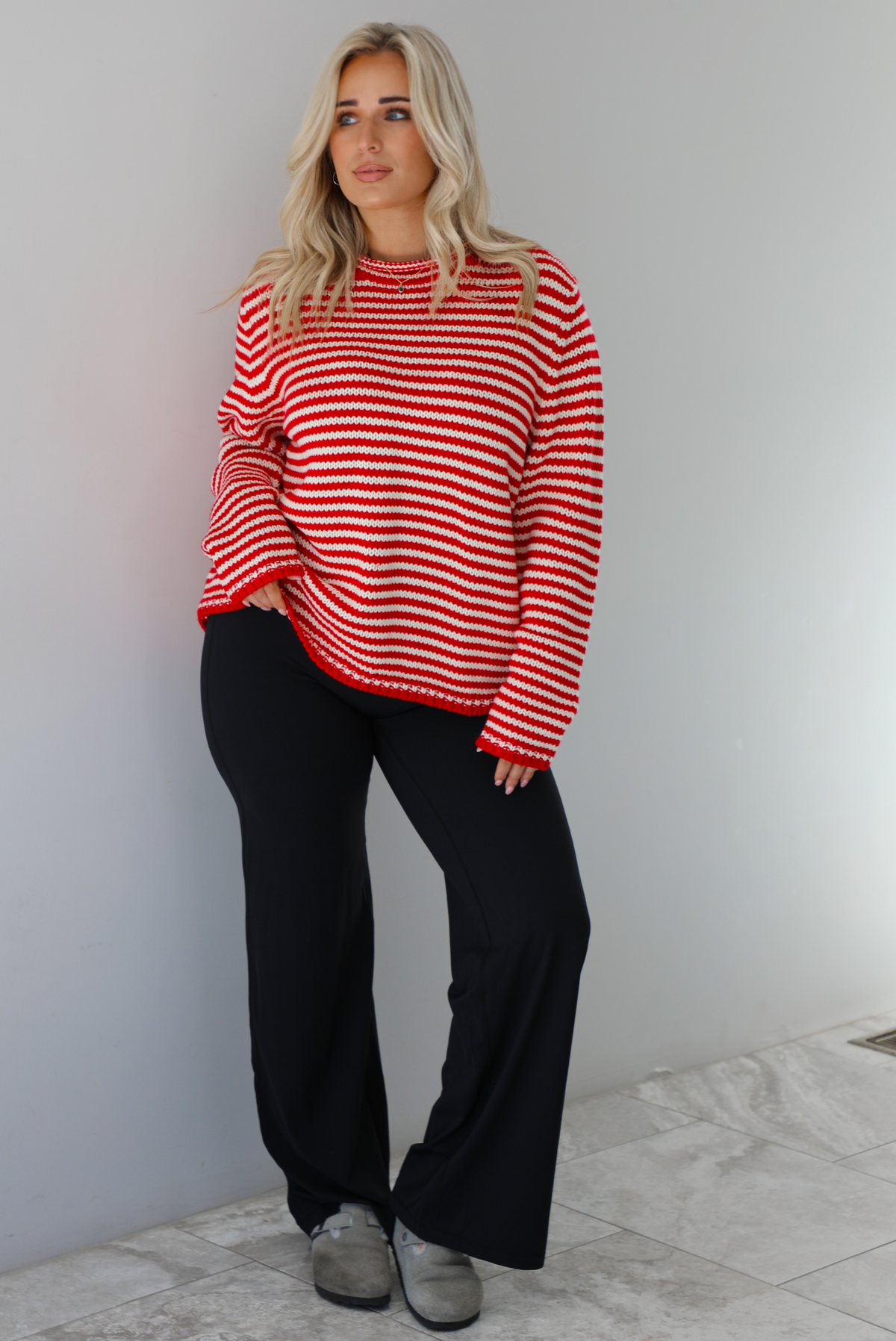 Woman wearing a white and red striped sweater that has a high neckline, an oversized fit, and is paired with black leggings. The photo is taken indoors against a white wall with white marble flooring. 