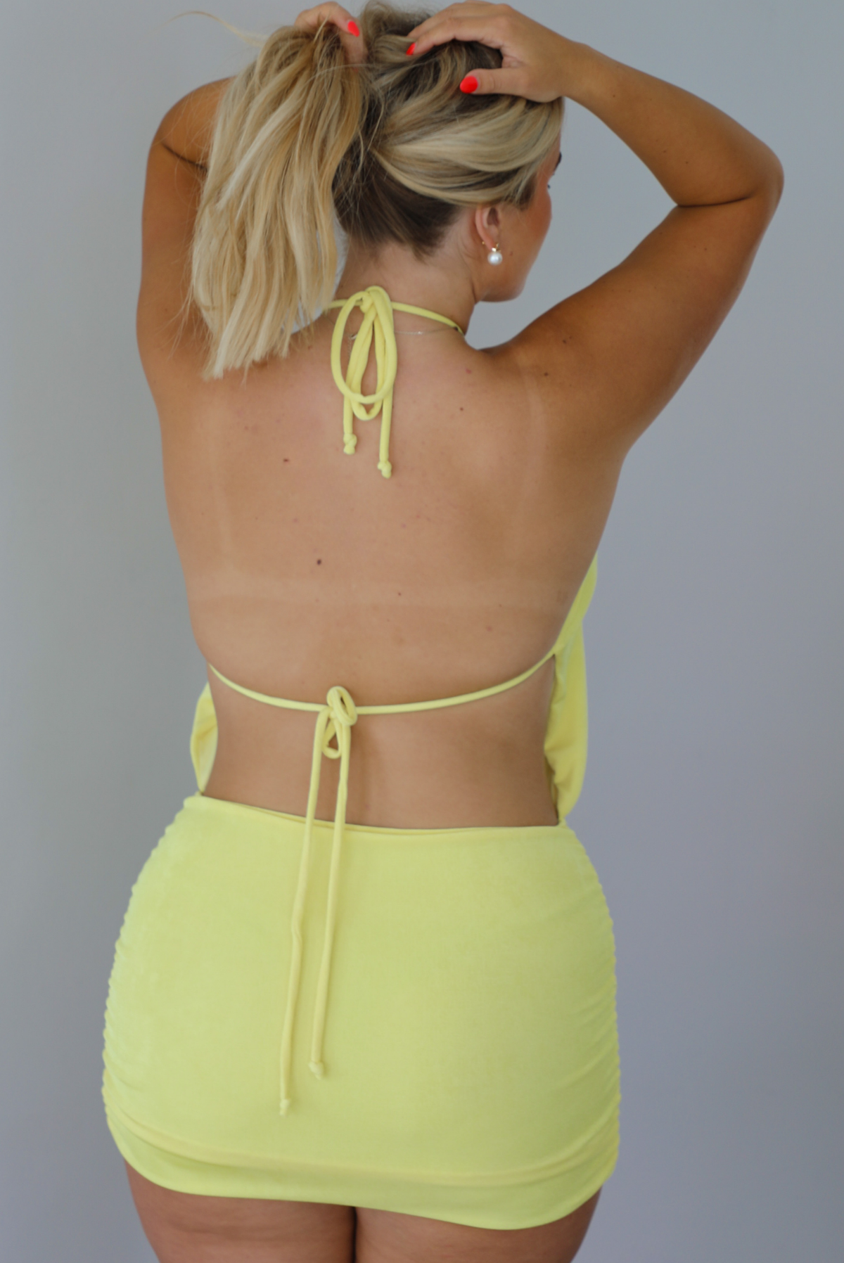 A blonde woman modeling a sleeveless, high-neck mini dress in a bright neon yellow with a slouched bodice and fitted skirt, indoors against a white wall. The photo shows the back of the dress, which has an open back design and a tie closure in the center of the back.