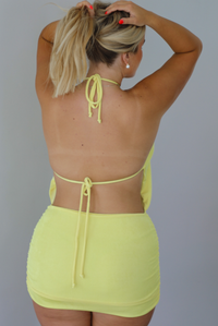 A blonde woman modeling a sleeveless, high-neck mini dress in a bright neon yellow with a slouched bodice and fitted skirt, indoors against a white wall. The photo shows the back of the dress, which has an open back design and a tie closure in the center of the back.
