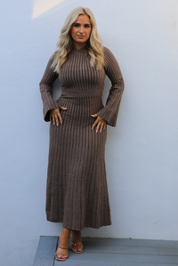 Woman wearing a taupe colored dress that has a vertical ribbed pattern throughout, a high neckline, fitted bodice, cinched waistline, long sleeves that flare slightly, and a maxi length skirt that flows at the bottom. The photo is taken outdoors against a white wall. 