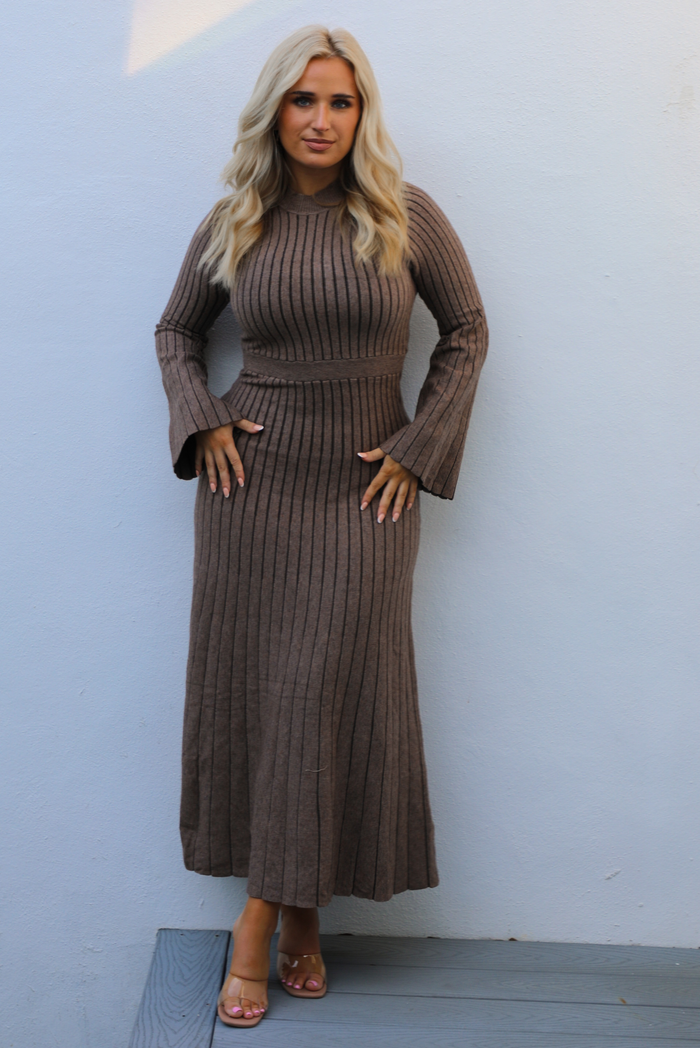Woman wearing a taupe colored dress that has a vertical ribbed pattern throughout, a high neckline, fitted bodice, cinched waistline, long sleeves that flare slightly, and a maxi length skirt that flows at the bottom. The photo is taken outdoors against a white wall. 