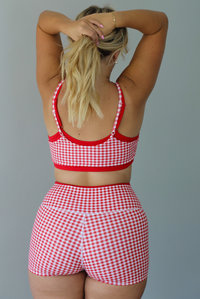 A woman modeling a red and white gingham two-piece athletic set featuring a sports bra and high waisted biker shorts with solid red trim indoors against a white wall. Th photo shows the back of the set, showing how the straps on the top are adjustable.