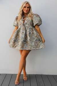 Woman model wearing a champagne and multi-colored floral pattern throughout. The dress has puffed sleeves, a v-neckline with tie details, and a baby doll cut that stops mid thigh on the 5'7" mode. The photo is taken outdoors against a white wall. 