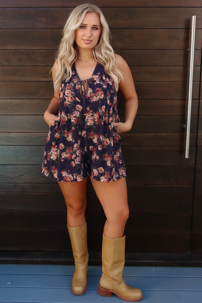 Woman model wearing a navy with pink and muted red floral print patterned denim romper that has a v-neckline with a tie closure in the center, related details along the bodice, functional pockets on either side, and a relaxed fit. It is paired with san colored boots that stop just under the knee on the model. The photo is taken outdoors against a dark brown wooden door.