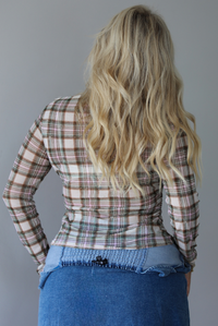 A woman shown from behind close up wearing a multi-colored plaid sheer long sleeved top that looks distressed, indoors against a white wall. 