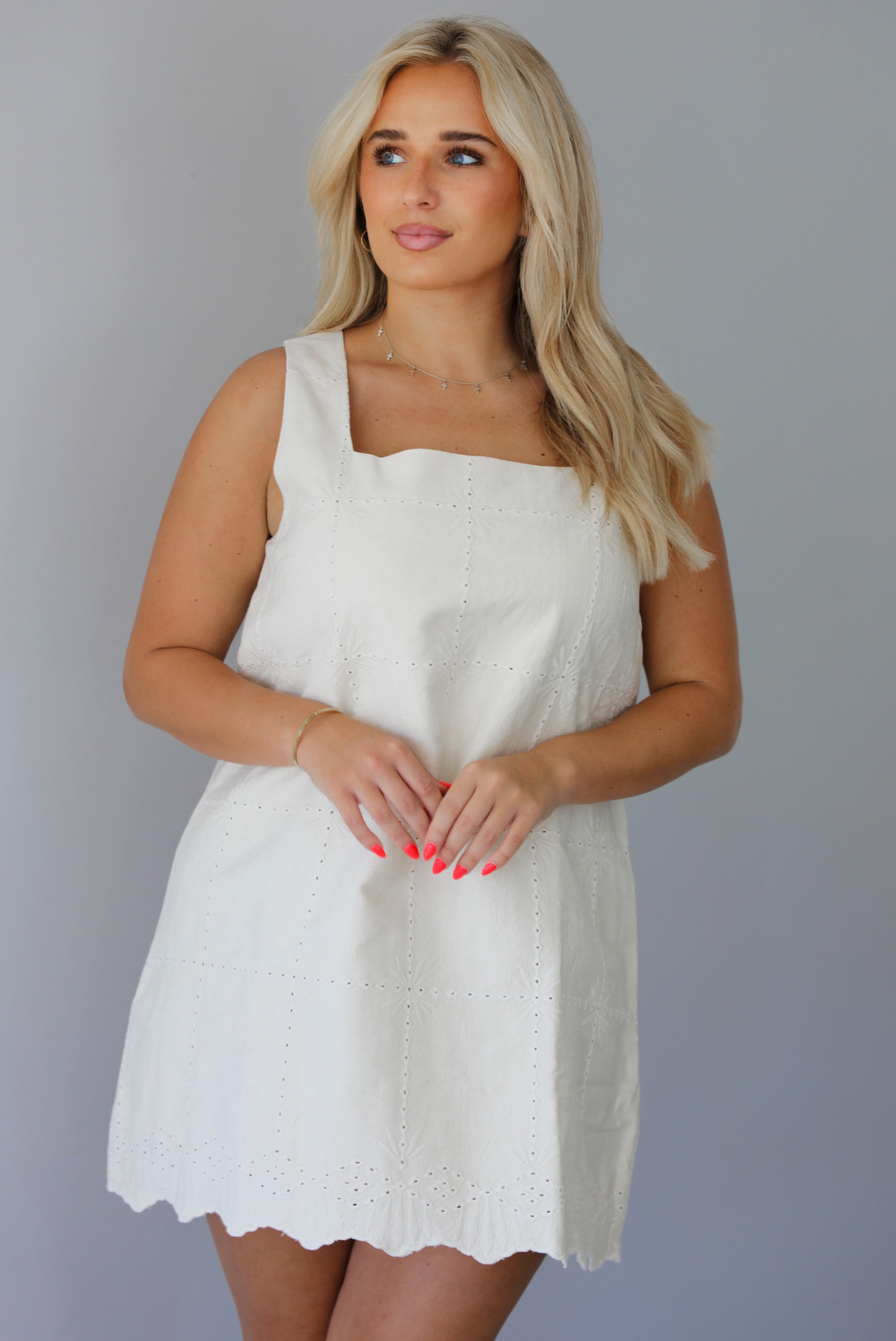 Woman wearing a sand colored mini dress with a square neckline, eyelet details throughout, and a scalloped hem indoors against a white wall.