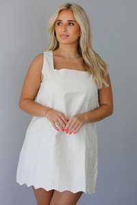 Woman wearing a sand colored mini dress with a square neckline, eyelet details throughout, and a scalloped hem indoors against a white wall.