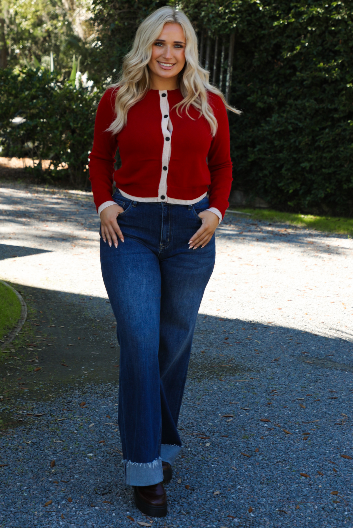 Woman model wearing a red cardigan with sand trim details and black button closures in the front with dark wash denim. The photo is taken outdoors on gravel with greenery in the back. 