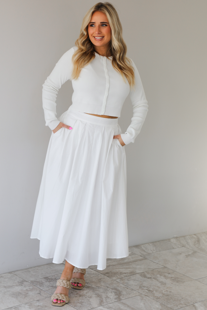 Woman wearing a white two piece set featuring a long sleeved cardigan and a high waisted midi length skirt. The photo is taken indoors against a white wall with white marble flooring. 