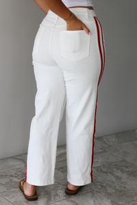 Woman wearing a white tank with white denim jeans with red stripes on either side  indoors against a white wall with white marble flooring. The photo shows a closeup of the back of the pants. 