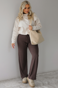 Woman wearing a beige high-neck, oversized sweater, high waisted brown pants, with a beige shoulder bag, and beige ugg boot shoes. The photo is taken indoors against a white wall with white marbled flooring. 