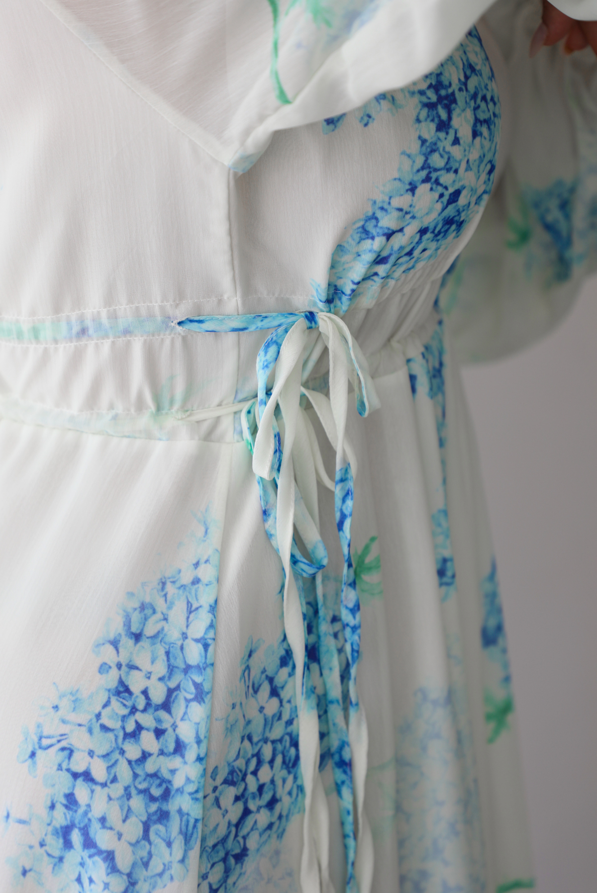 Closeup of a woman modeling a white maxi dress with a blue floral print and a sheer, flowing overlay, focusing on the side of the bodice of the dress with its cinched waistline, standing indoors against a white wall. 