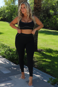 A woman modeling a matching black activewear set consisting of a sports bra and high-waisted leggings, standing outdoors on a paved path with a green lawn and trees in the background.