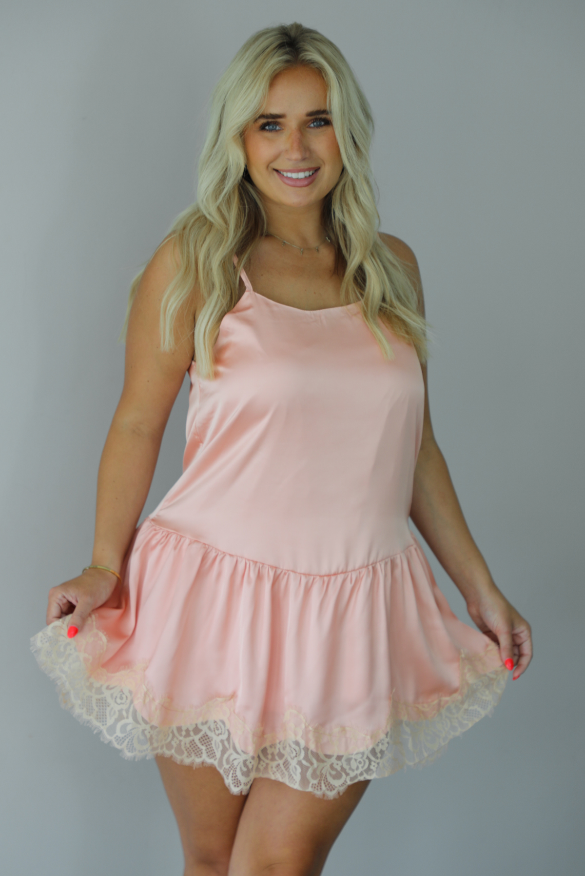 Woman wearing a peach colored satin and lace mini dress indoors against a white wall. The photo shows a closeup of the dress, showing how it's entirely satin with a lace trim design. 