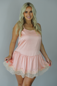 Woman wearing a peach colored satin and lace mini dress indoors against a white wall. The photo shows a closeup of the dress, showing how it's entirely satin with a lace trim design. 