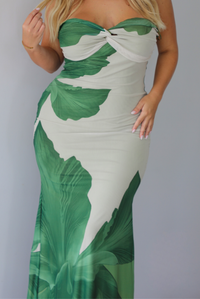 A closeup of a blonde woman models a strapless, bodycon maxi dress with a green tropical leaf print on a light beige background, standing indoors against a white wall.