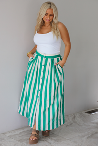A woman wearing a white tank top and a maxi length cream and green striped skirt,  which has a high waistline, functional pockets on either side, functional button closures in the front, and has a flowy fit, standing indoors against a white wall with white marble flooring. 