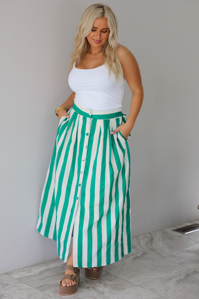 A woman wearing a white tank top and a maxi length cream and green striped skirt,  which has a high waistline, functional pockets on either side, functional button closures in the front, and has a flowy fit, standing indoors against a white wall with white marble flooring. 