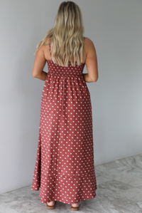 Woman wearing a spaghetti strap, maxi length dress that has a rust color throughout with a white polka dot pattern. The photo is taken indoors against a white wall with white marble flooring and shows the back of the dress, which has a smocked back and adjustable straps. 